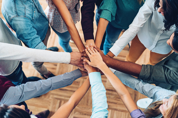 Eleven arms of various skin hues and wearing shirt sleeves of various lengths extend into the center of a circle, where they stack on top of one another in a gesture of camaraderie.
