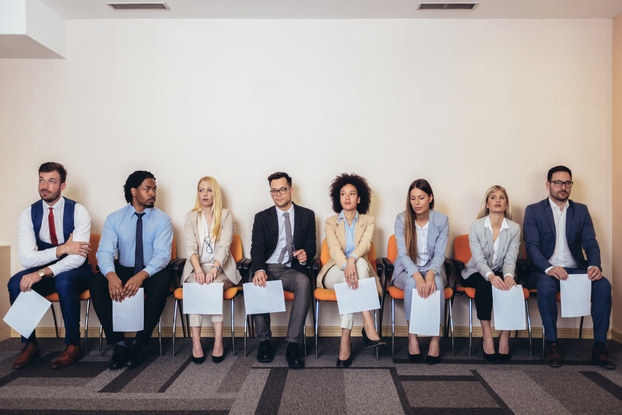 A row of people of many ethnicities and genders sit in chairs. They are all dressed in business casual wear and each person holds a piece of paper.