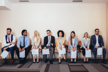  A row of people of many ethnicities and genders sit in chairs. They are all dressed in business casual wear and each person holds a piece of paper. 