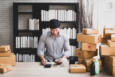 A man stands behind a counter piled with cardboard boxes. The boxes are taped closed and many are marked with red circles containing either the letter A or the letter C. The man is looking down the smartphone held in his hand and a calculator on the desk. 