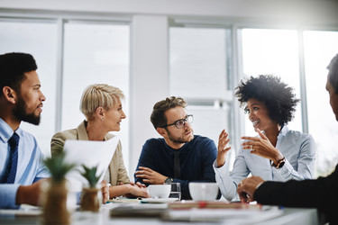  A group of five people sit around a conference table. Everyone's attention is on the fourth person from the left, a woman who is excitedly talking and gesturing with her hands. 
