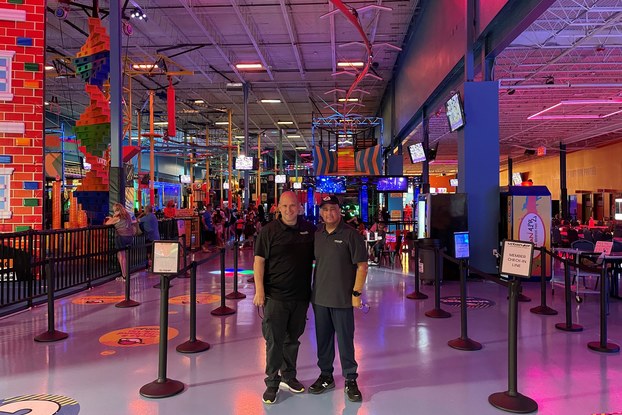 Two men stand next to each other inside an entertainment facility with crowd-control barriers and neon lights