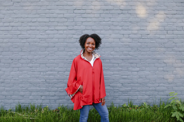 Woman smiles as she stands in front of a white brick wall and grass, wearing a dark orange rain jacket.