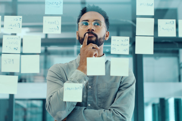 A man stands with one hand on his chin, considering the transparent glass between him and the viewer. On the glass are several post-it notes, some with writing on them.
