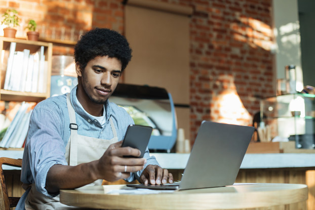 Man in apron sits at table and works on smartphone and laptop in interior of loft coffee shop.