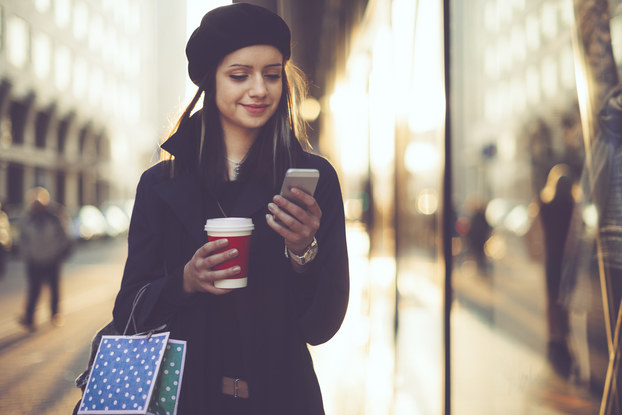 Woman holding shopping bags and coffee and looking at phone.