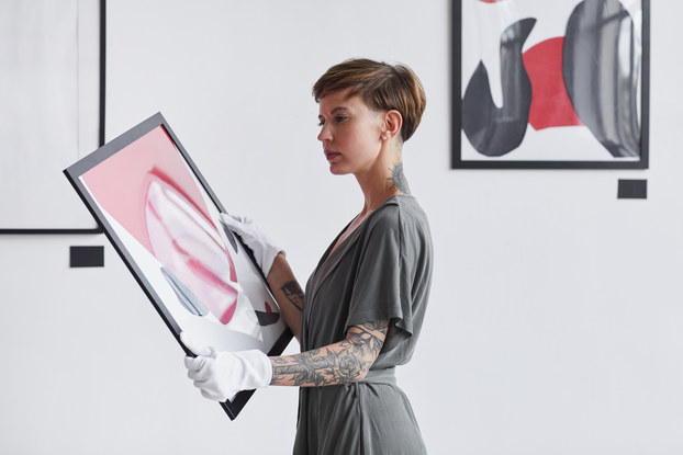 A woman wearing white gloves stands in a gallery holding a framed painting, looking thoughtful. The woman wears a gray dress and has short hair and a sleeve tattoo on her left arm. She examines an abstract painting made of pink, white and gray slashes and strokes.