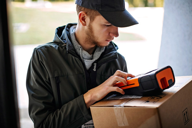 A young man wearing a windbreaker and a baseball cap examines a scanning device, which he balances on top of a package in a brown cardboard box.
