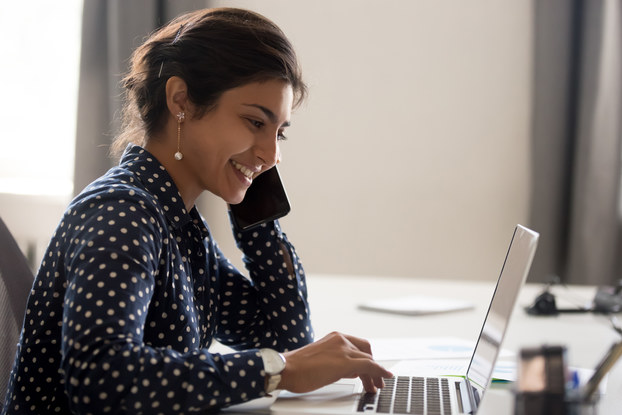 A woman wearing a blue-and-white polka-dotted shirt looks at an open laptop with a smile.