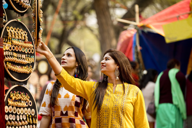 Two women with long dark hair look at hanging displays of large handmade earrings, most of them dangling and gold or copper. The earrings are stuck in circular boards hanging from an unseen structure. In the background are trees and part of a market stall around which other patrons clusters.