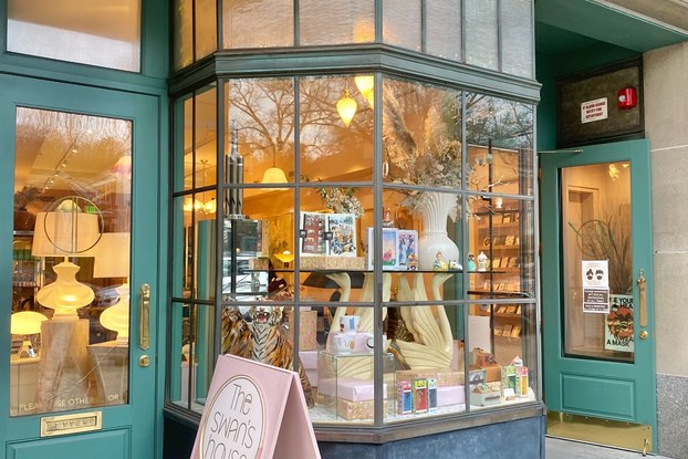 Portrait of a shop's exterior with two green doors and a bay window, the shop's sign sits out front on the sidewalk.