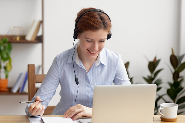 A woman wearing a button-up shirt and headphones is sitting at a table and looking at a laptop with a smile. She holds a pen over a notepad with her right hand.