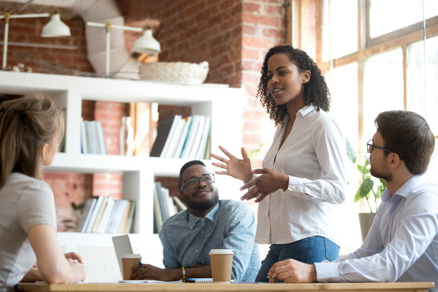 A young woman stands in front of a table in a brick-walled office space with a large floor-to-ceiling window at her back. She talks and gestures with her hands. Around the table sit three of the woman's colleagues, who look up at her in interest.