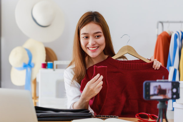 A woman holds up a red sweater on a hanger and looks in the direction of a smartphone on a tripod in the foreground. The smartphone is positioned horizontally and the image of the woman on the sweater can be seen out-of-focus on its touchscreen. In the background are more sweaters on hangers hanging on a rack and another rack holding wide-brimmed sunhats.