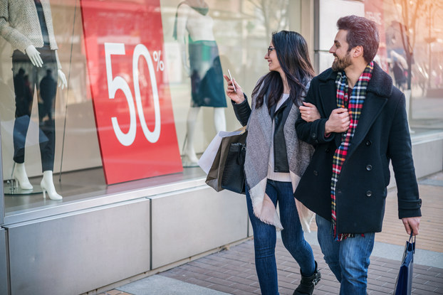 A couple walks arm-in-arm past a store window. In the window stand two mannequins, one in dark pants and a tweed jacket and the other in a dark skirt, white blouse and black scarf. Between the mannequins is a huge red sign that says "50%" in white text.