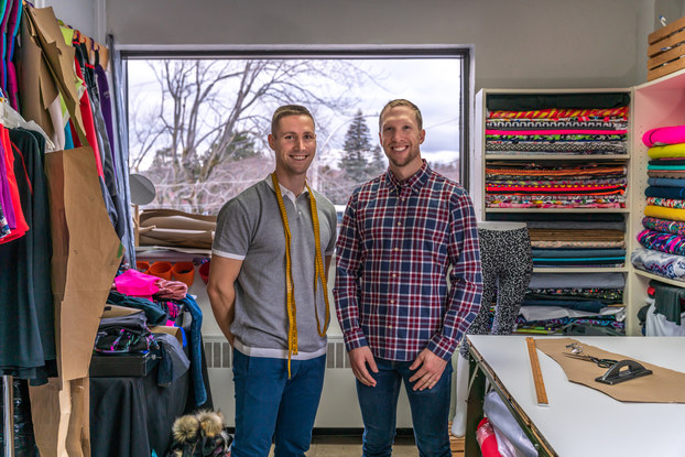 Two men stand smiling in a fabric store among shelves and racks of various brightly colored swatches of cloth. The man on the left stands with his hands behind his back and wears jeans and a gray-and-white polo shirt. He has bright yellow measuring tape draped around his neck. The man on the right wears jeans and a long-sleeved flannel shirt. Near him in the foreground is a table with a cut-out piece of khaki fabric, a yardstick and other measuring tools on top.