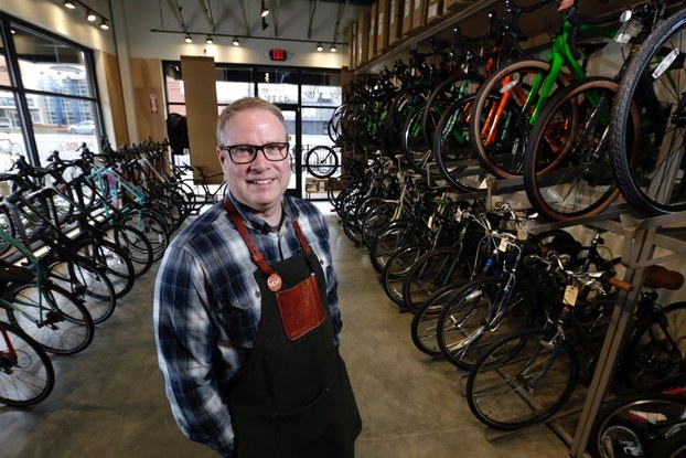 A smiling man wearing glasses, a plaid shirt and an apron stands in a bike shop with rows of bikes behind him.