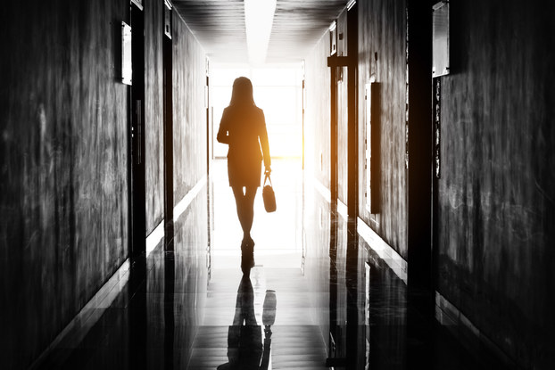 A silhouette of a woman wearing a knee-length dress and holding a bag in one hand walks down a dark office hallway lined with doors toward a golden light.