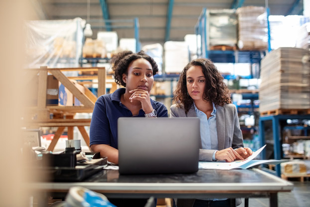 Two women stand at a table in the middle of a warehouse and look curiously at a laptop. IN the background are shelves filled with pallets of goods wrapped in plastic.