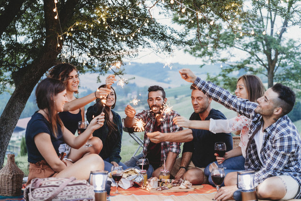 A group of seven friends sit on an orange picnic blanket outside, with a scenic valley in the background and trees strung with lights overhead. In front of the group are glasses of wine, a couple of lanterns, a charcuterie board, and mason jars filled with a golden beverage. Each member of the group holds up a lit sparkler.