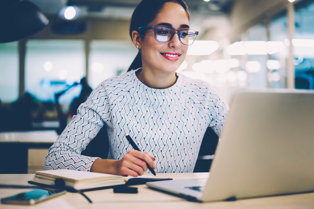 Young business woman is pleased with information she sees on her laptop and takes notes.