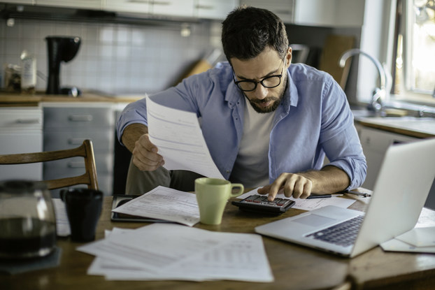 Man sits at kitchen table with laptop, papers, and calculator. He calculates information while drinking coffee.