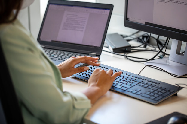An over-the-shoulder shot of a person with long dark hair typing on a keyboard. The keyboard is connected to a computer monitor that displays a text document. To the person's left is a laptop with a monitor also displaying a text document.