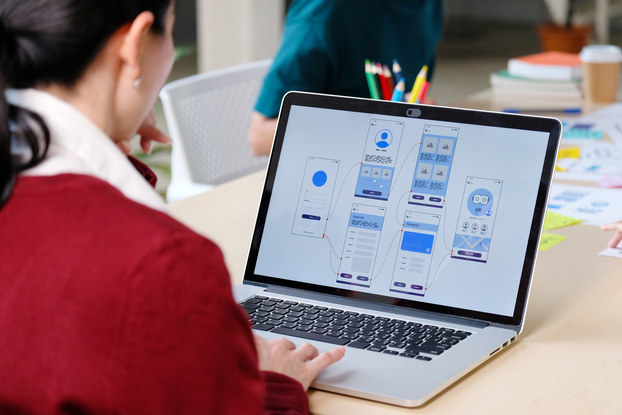 An over-the-shoulder shot of a woman wearing a red sweater. In front of her on a table is a laptop with a monitor showing a series of connected boxes with various graphs and pictures in shades of blue and gray.