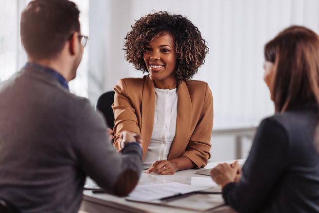 A woman in a brown blazer sits facing the camera. Across a table from her are another woman and a man who both face away from the camera. The woman facing the camera shakes the man's hand with a smile.