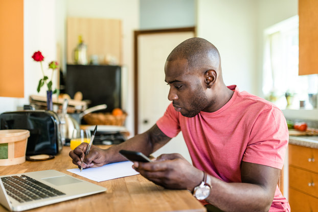 Man works at kitchen table with calculator, pen, notebook, and laptop.
