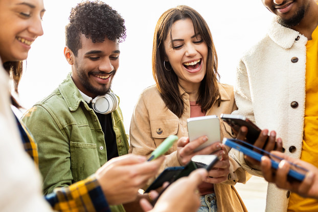 A diverse group of young people stand in a tight circle and hold their smartphones in front of them. Their eyes are on their phones and they're all smiling.