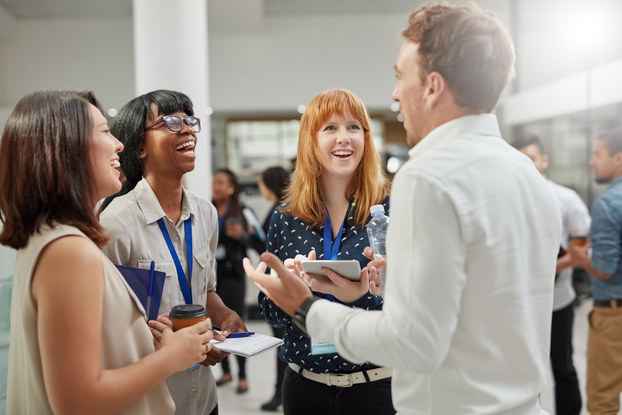 A group of people (three women and one man) stand in a circle and laugh. The two women in the middle of the photo are wearing blue lanyards; a card at the end of one of the lanyards reads "CONFERENCE." In the background, other groups of people, some of whom are also wearing blue lanyards, can be seen.