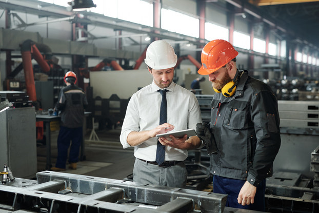 Two men wearing hard hats stand side by side in a warehouse filled with various machines and steel tables. Both men look at an electronic tablet held by the man on the left.
