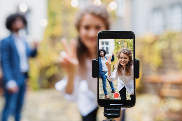 In the foreground and slightly right of center, a smartphone in a tripod holder shows a woman and, a few feet behind her, a man. Both of them are standing outside in front of a wall draped in ivy, making peace signs at the camera. In the background out of focus, the woman and man pose for the camera.