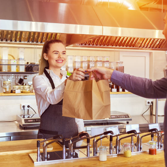 A woman in an apron stands behind a wooden counter and hands two paper bags to a customer.