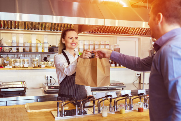 A woman in an apron stands behind a wooden counter and hands two paper bags to a customer.