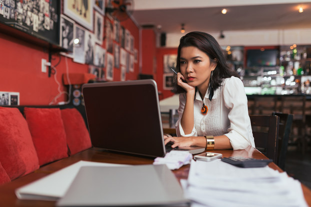 A woman uses her laptop at a paper-strewn table in an eclectically decorated restaurant. She looks thoughtful.