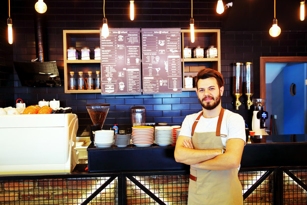 A man wearing an apron stands in front of a coffee shop counter, behind which is mounted a menu.