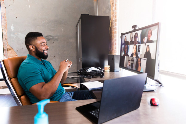 A man sits at a desk facing a large monitor with a webcam. On the monitor are video feeds of several other people in a meeting.