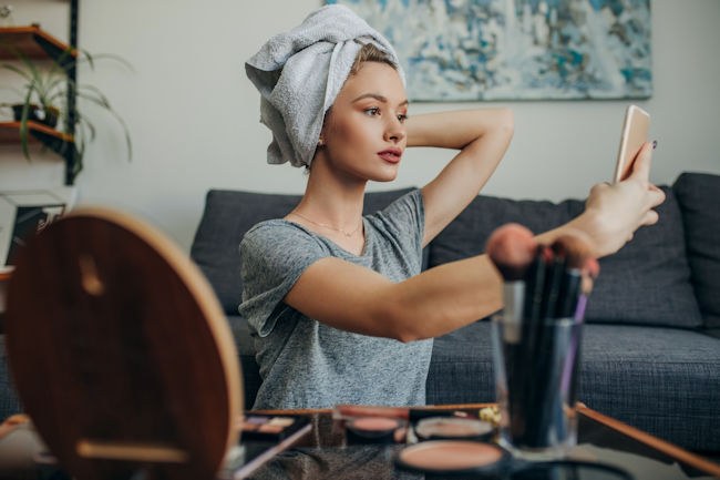 A woman with a towel on her head who has just applied her makeup takes a selfie with her smartphone.