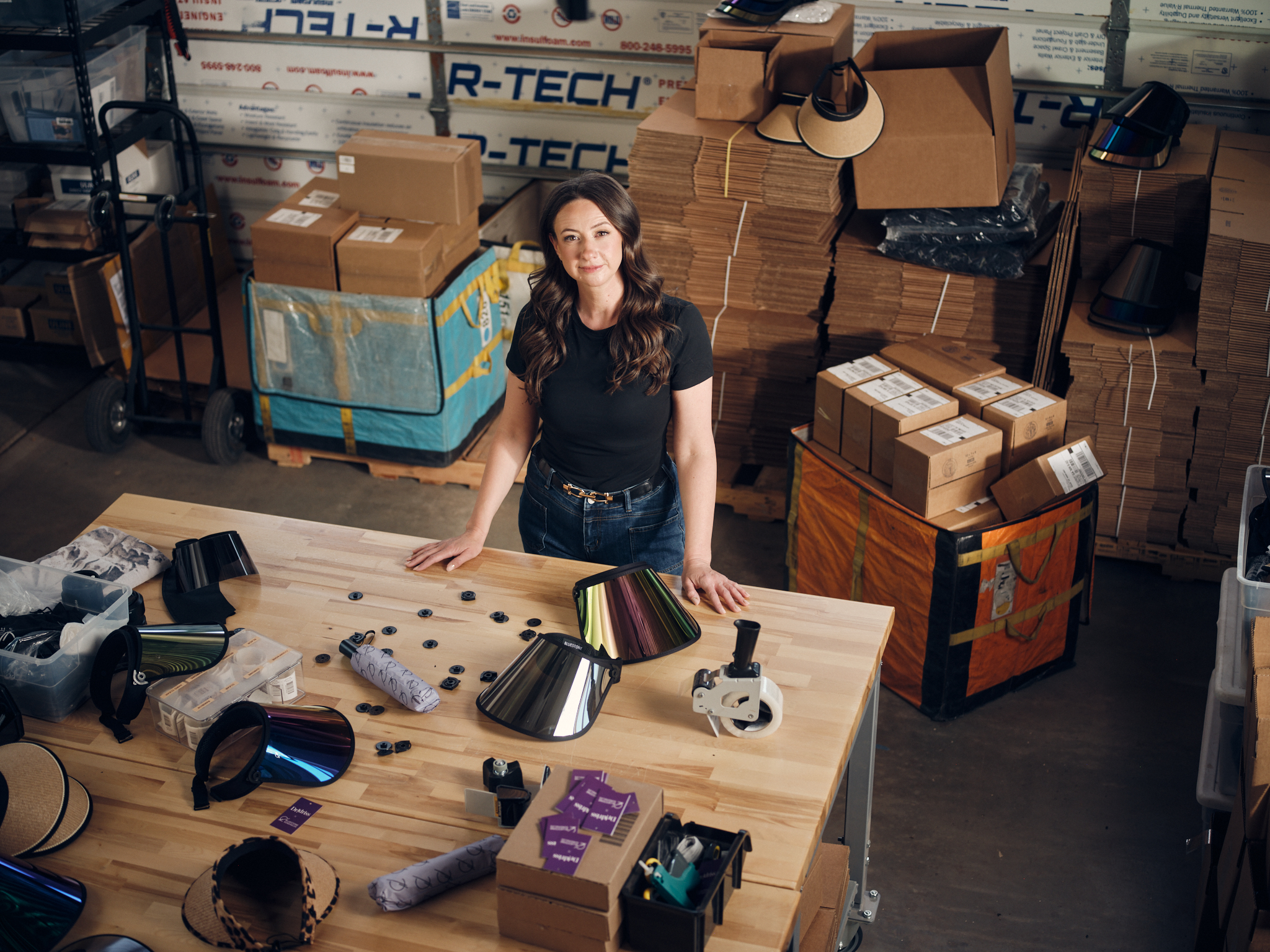 A shot from above of Jen Podany, Founder of Bluestone Sunshields, standing at a wooden work table. Jen has long wavy dark hair and is wearing a black T-shirt and dark wash jeans. In the background are stacks of cardboard boxes.