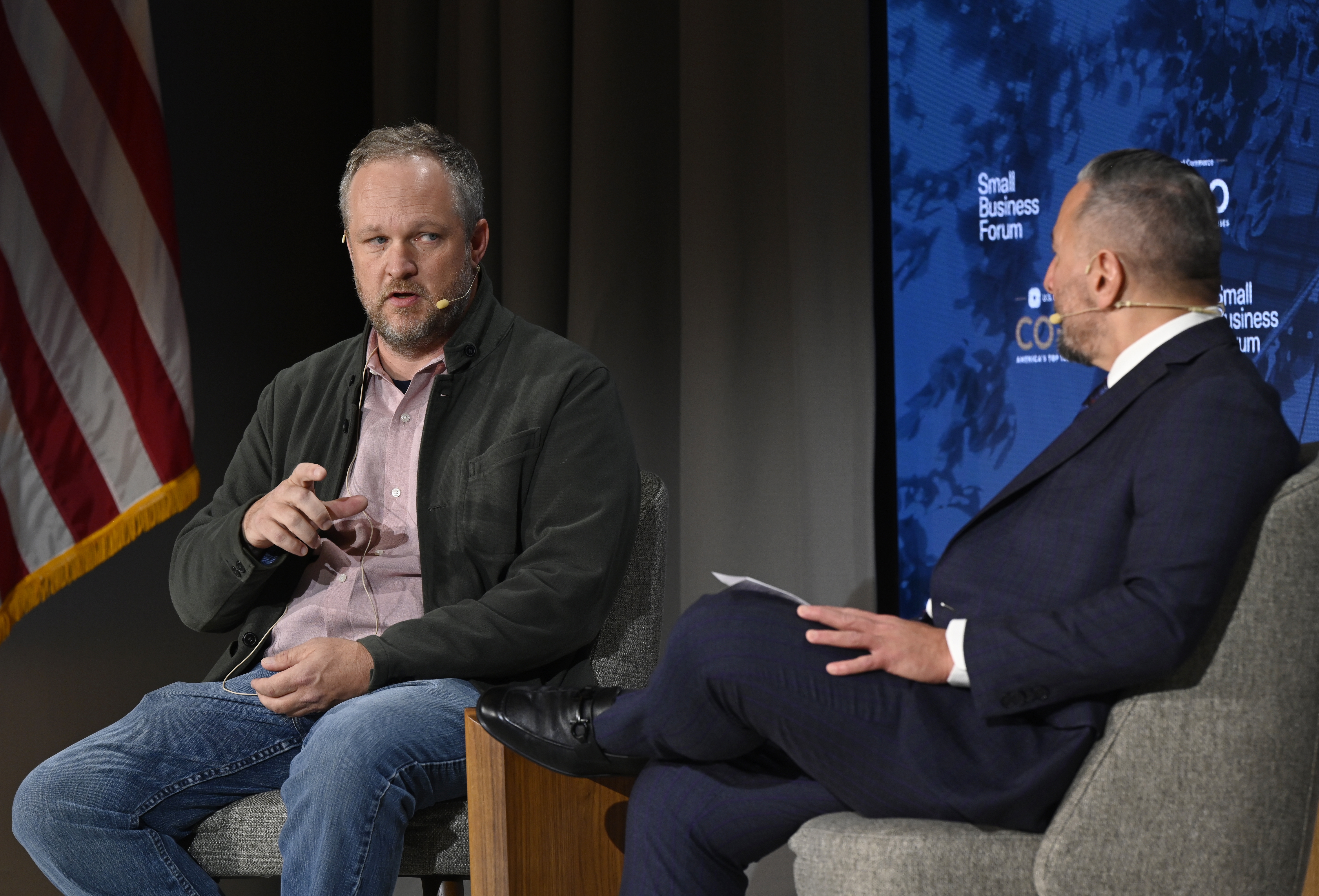 Two individuals engage in a discussion on a stage at the Small Business Forum, with an American flag visible in the background.