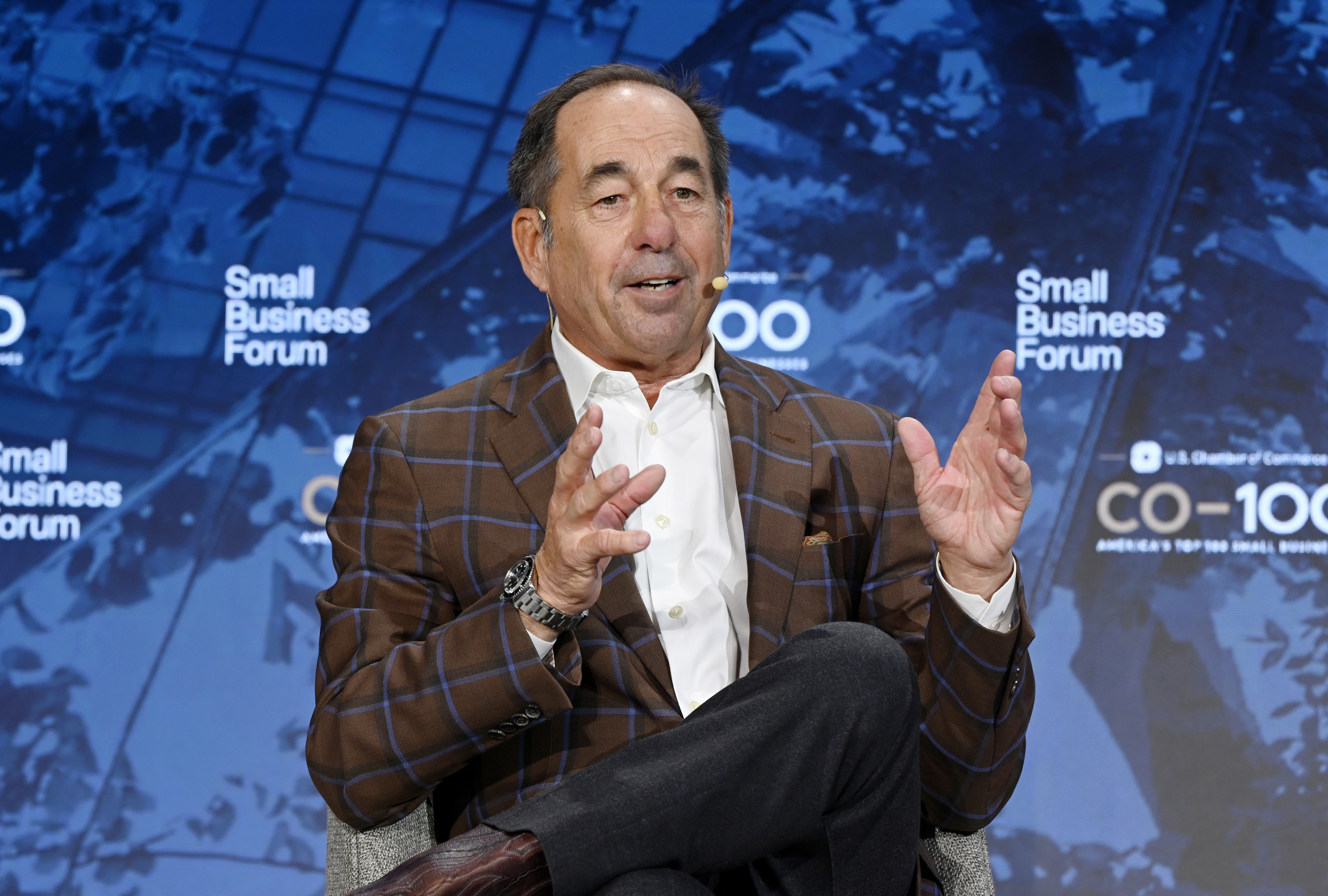 A person in a plaid suit speaks at a Small Business Forum event, gesturing with both hands while seated on a stage.