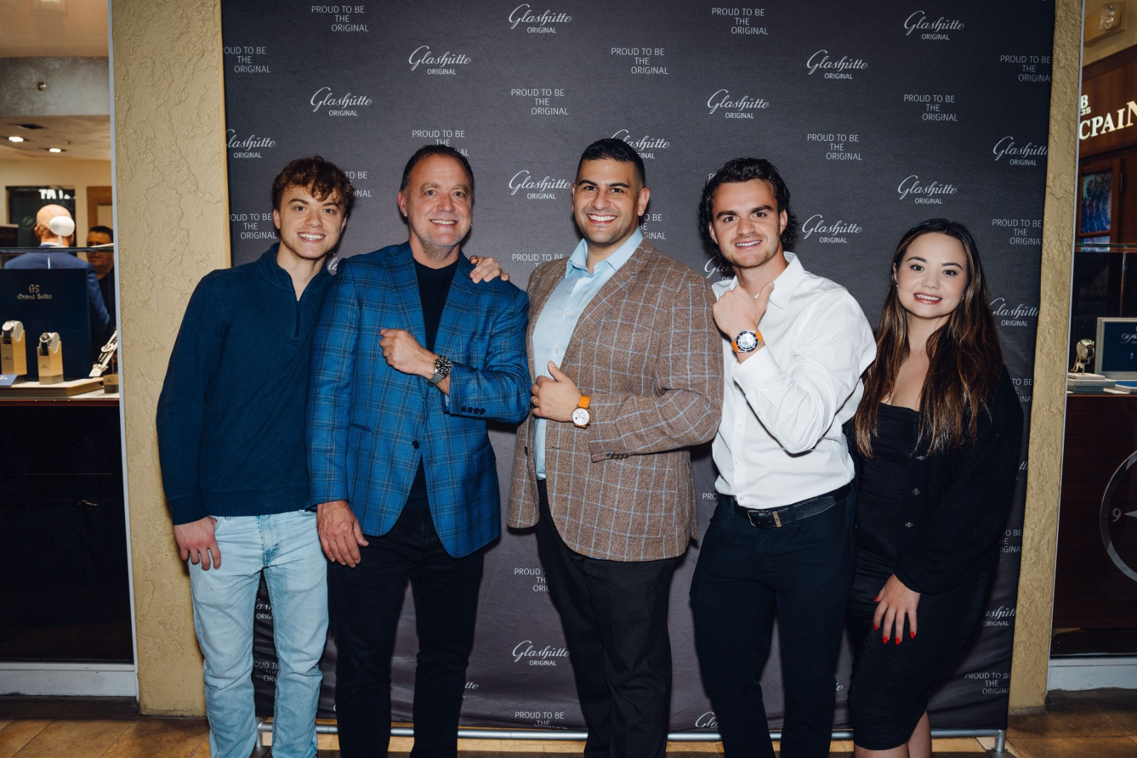 A family of five people stands together in front of a black backdrop. The three adult men in the center have their arms raised, showing off the luxury watches on their wrists. On the ends of the group are a young man and a young woman.
