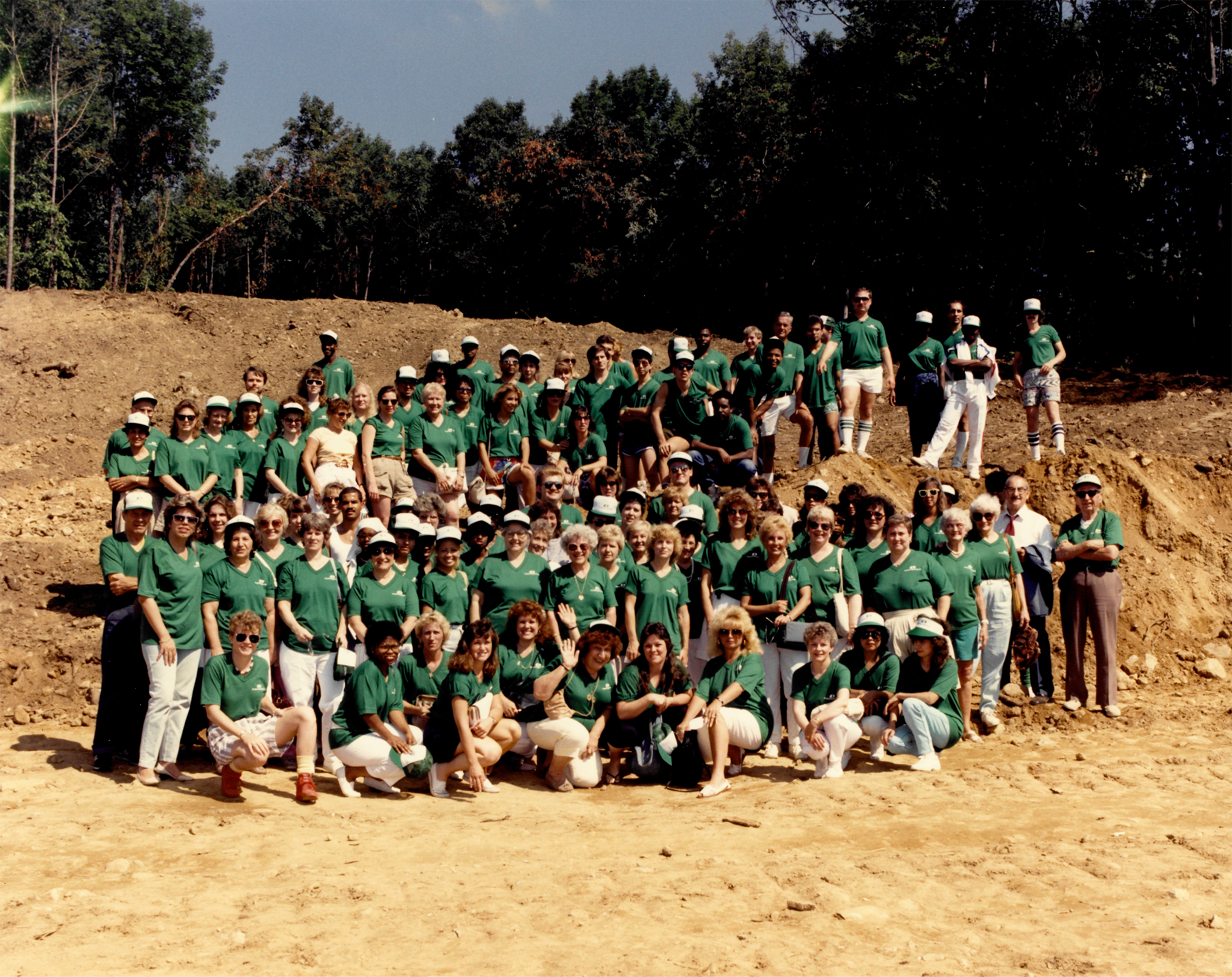 A wide shot from the 1980s, showing the Gallery Collection company team (about 75 people) posing for a photo outside. Every member of the team is wearing a green shirt and most are wearing white pants and white baseball caps.