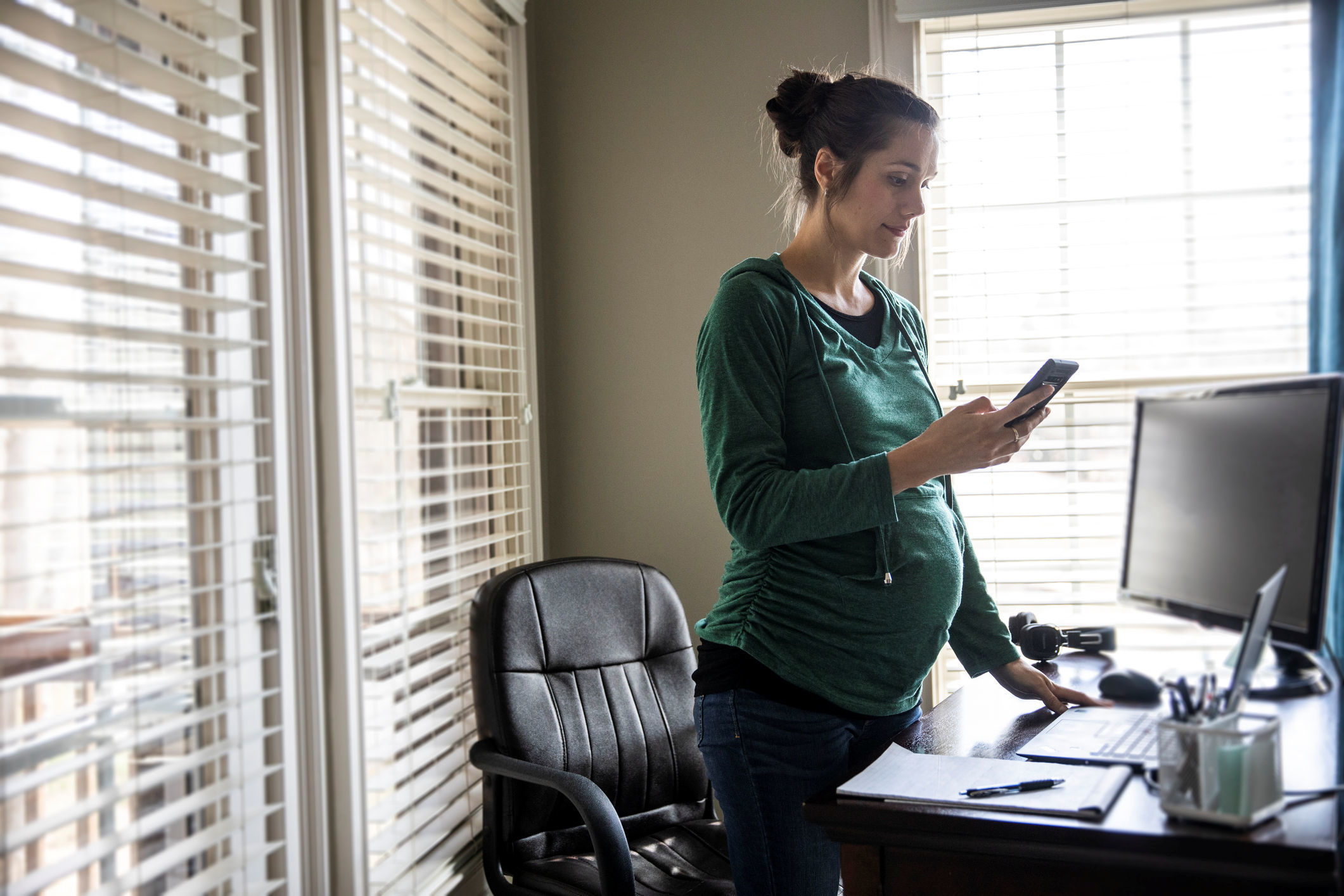 A young pregnant woman sands before her desk. She is holding her phone in her right hand and reading a message on her phone.