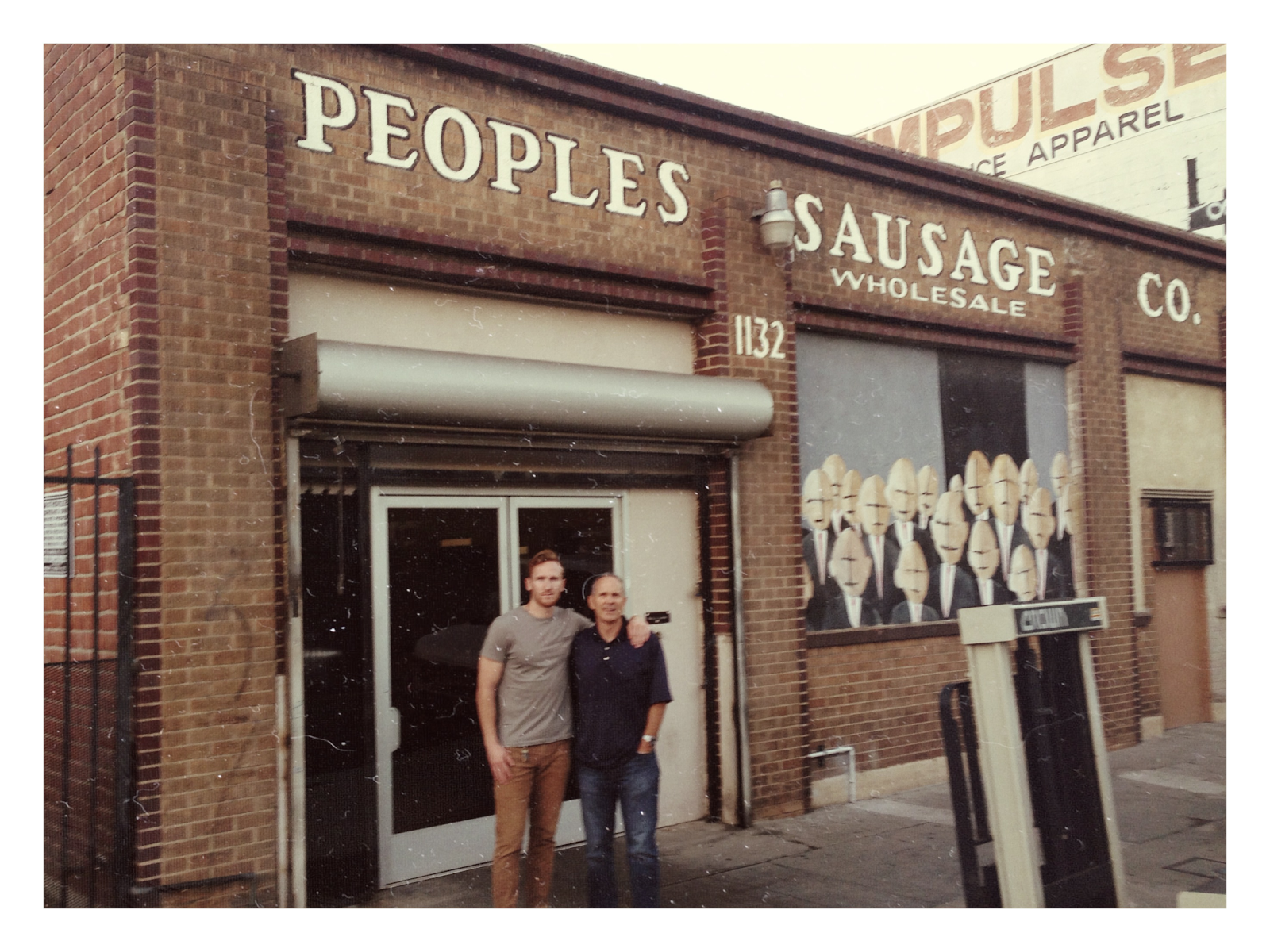 Two men standing in front a brick building with "PEOPLES SAUSAGE CO." written in white along the top of it. The men are Mark and Brian Bianchetti, third- and fourth-generation owners of People's Choice Beef Jerky.