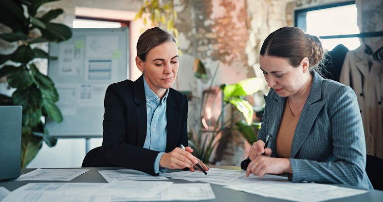 Two women are seated at a table and review documents. One woman uses a pen to point out something to the other woman.