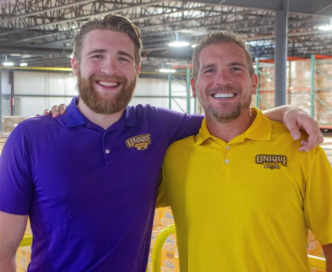 Justin Spannuth, COO of Unique Snacks, and his son Tristan, standing in a warehouse with their arms around each other's shoulders. Both men are wearing polo shirts with the Unique Snacks logo on the right side of the shirt.