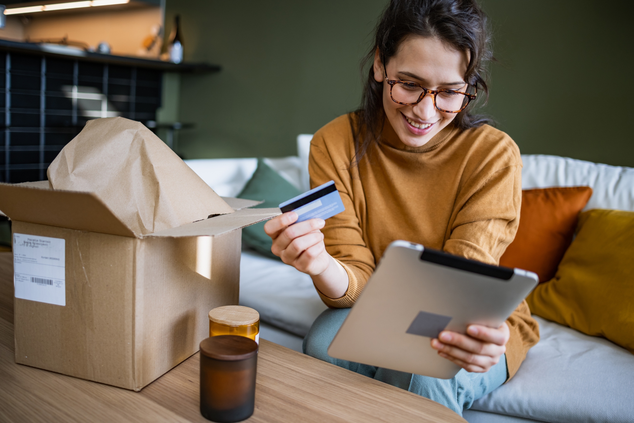A woman sitting on a sofa, holding a credit card and a tablet. She's looking at the tablet with a smile. On the coffee table in front of her are a couple of candles in glass jars and an open cardboard box with an address label on it.